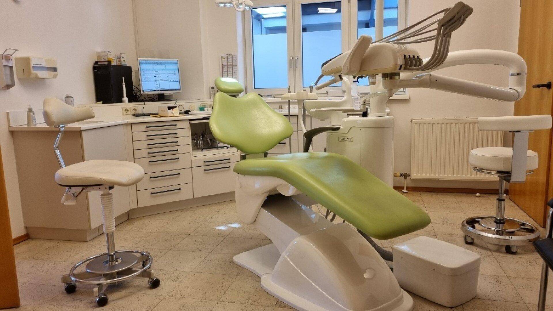 Behandlungszimmer mit grünem Behandlungsstuhl, weißen Schränken und medizinischen Geräten, ergonomisch gestaltet.