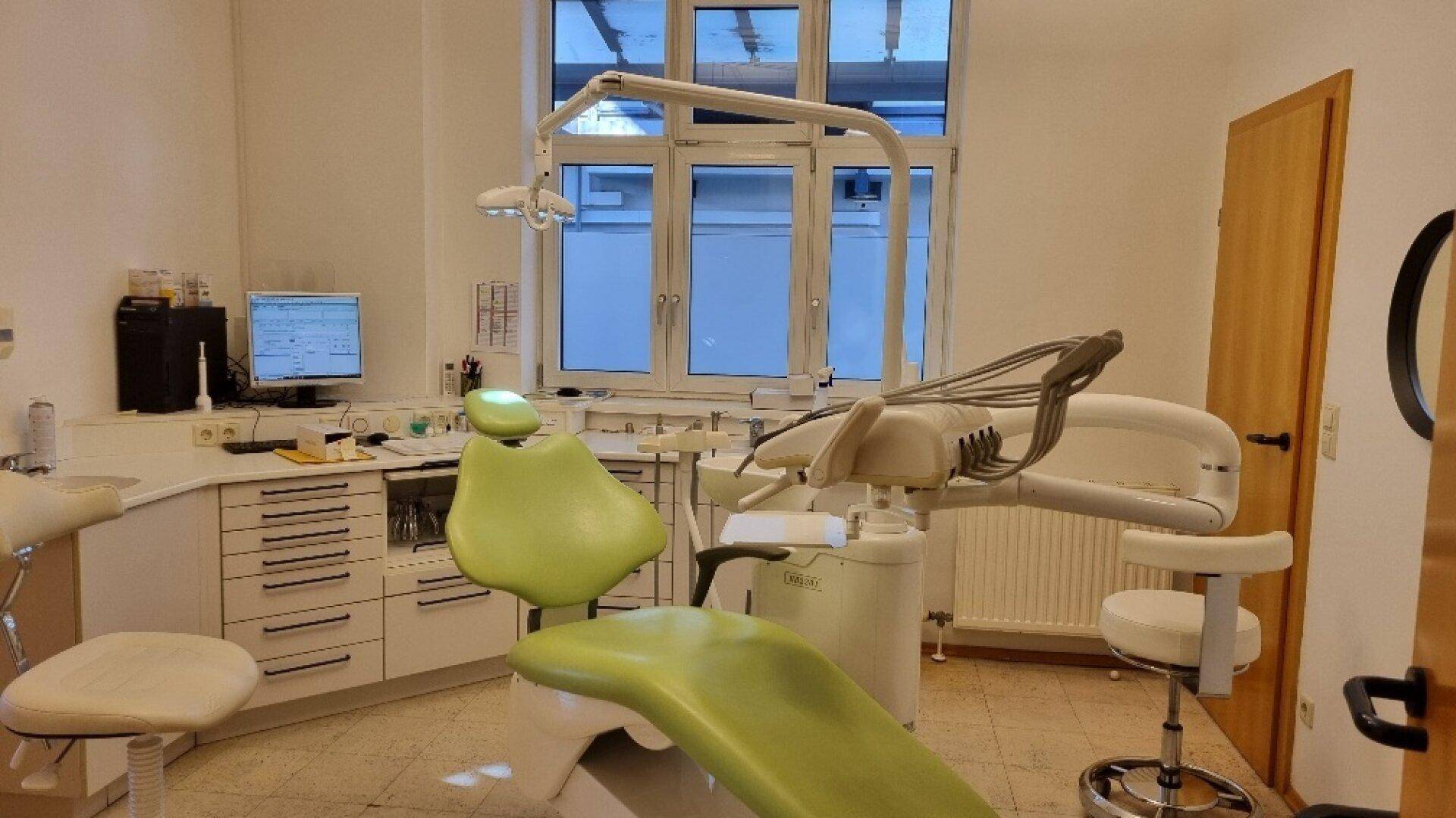 Behandlungszimmer mit zahnmedizinischer Einheit, Fenster und Heizkörper, sauber und funktional für Patienten.