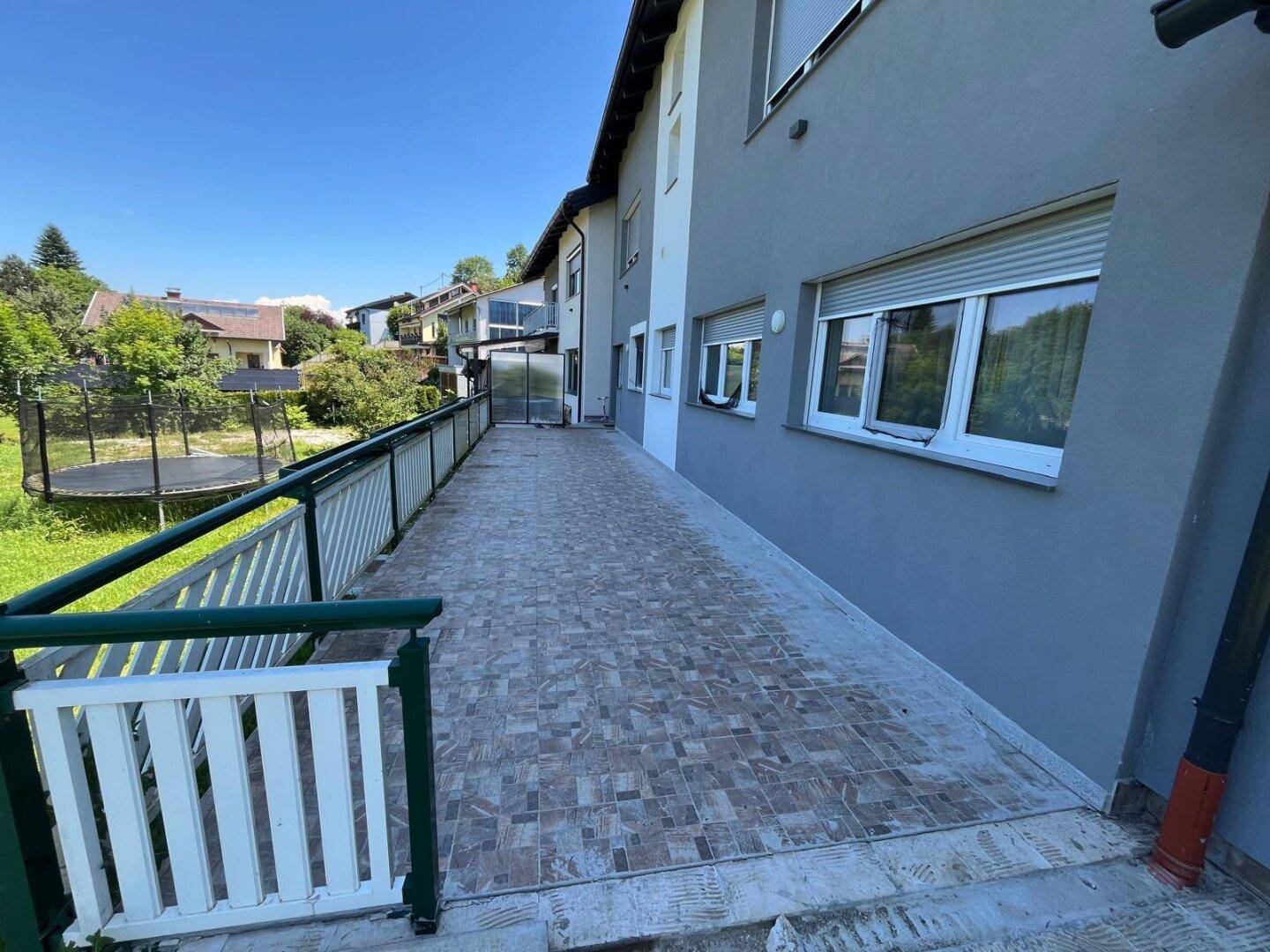 Geräumige Außenterrasse mit Fliesenboden und Blick auf den Garten mit Trampolin.