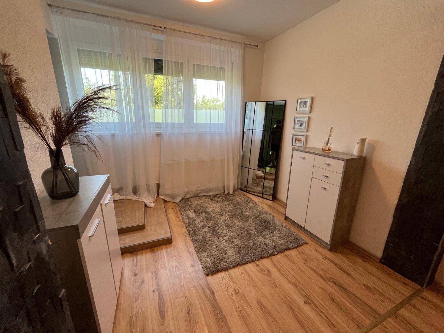 Heller Raum mit Holzfußboden, großem Fenster und modernem Spiegel, der viel Tageslicht hereinlässt.