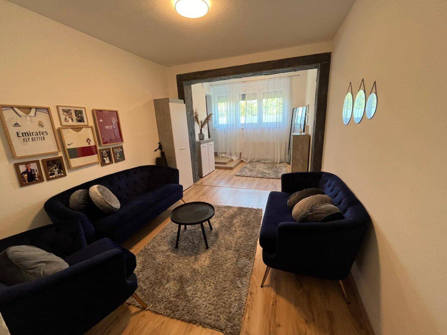 Stilvolles Wohnzimmer mit zwei blauen Sofas und Couchtisch, offen zu einem hellen Raum mit großem Fenster.