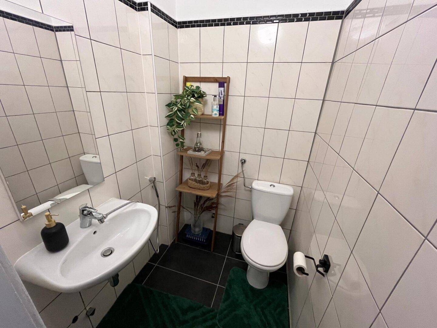Kleines Gäste-WC mit Waschbecken und Toilette, gefliest in Weiß mit schwarzem Akzentstreifen.