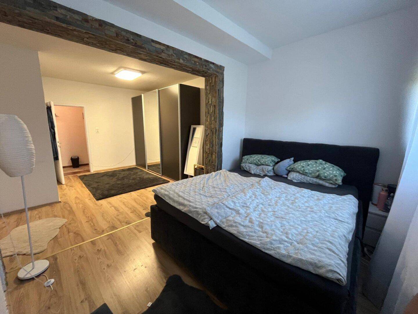 Gemütliches Schlafzimmer mit dunklem Bett, Holzfußboden und großem Spiegelschrank, verbunden durch einen Steinbogen.