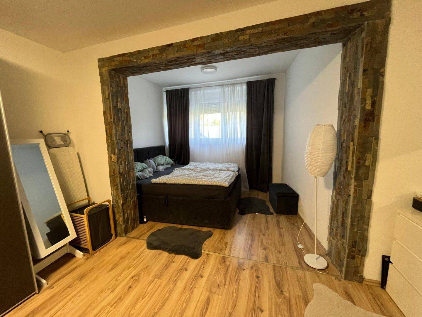 Ruhiges Schlafzimmer mit dunklem Bett und Holzfußboden, durch einen Steinbogen vom Hauptraum getrennt.
