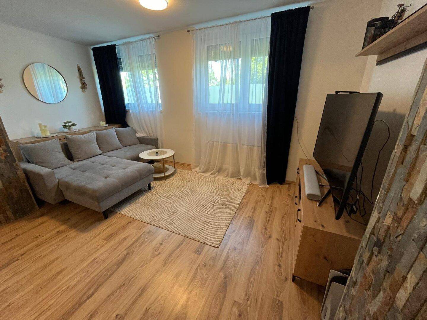 Gemütliches Wohnzimmer mit grauem Ecksofa, Holzboden und zwei großen Fenstern für viel Tageslicht.
