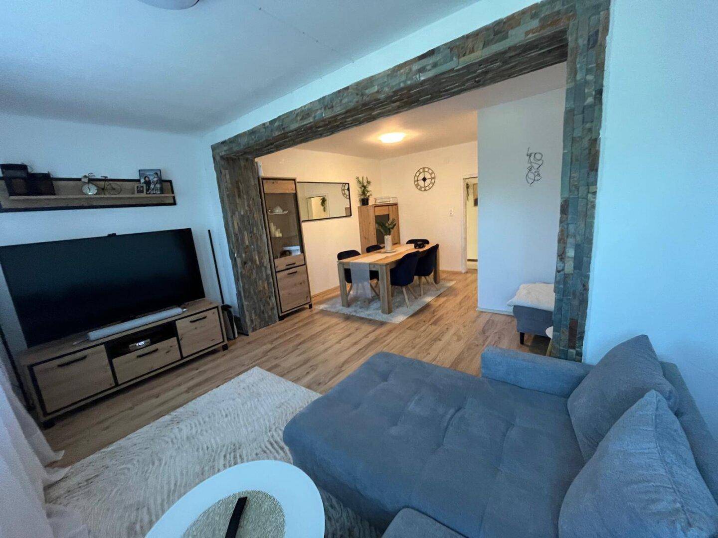 Offener Wohnbereich mit bequemem Sofa und Blick auf den Essbereich, verbunden durch einen dekorativen Steinbogen.