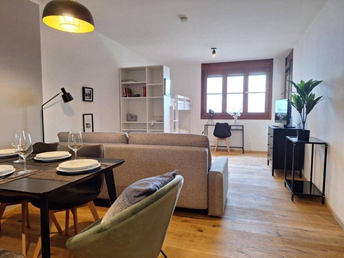 Heller Wohn-Essbereich mit Holzboden, Esstisch und Sofa, ergänzt durch ein Etagenbett im Hintergrund.