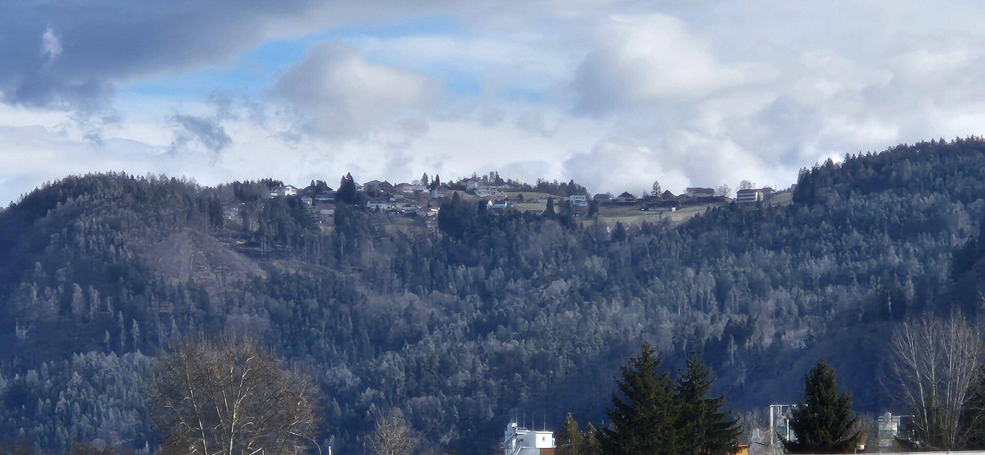 Blick auf eine Siedlung auf einem bewaldeten Hügel unter einem weiten Himmel.