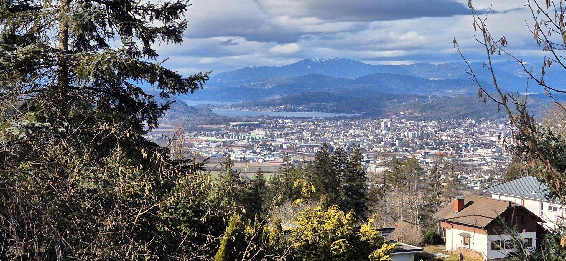 Weitblick über die Stadt Klagenfurt am Wörthersee mit See und Bergpanorama, umrahmt von Bäumen.
