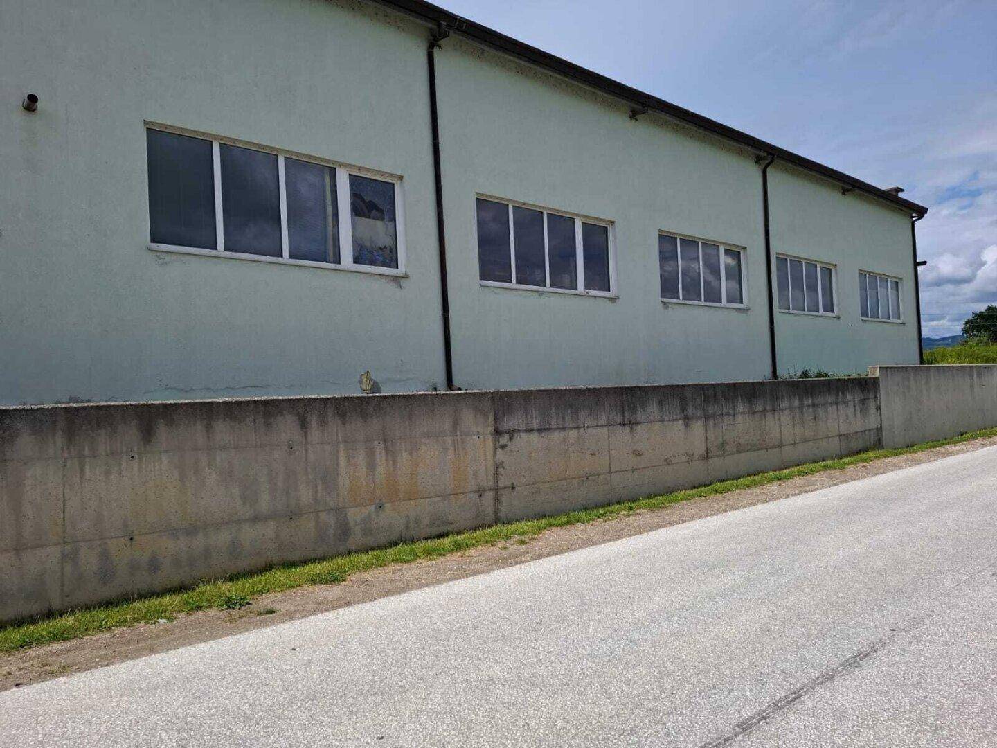 Außenansicht eines hellgrünen Industriegebäudes mit mehreren Fenstern und einer Betonmauer entlang der Straße.