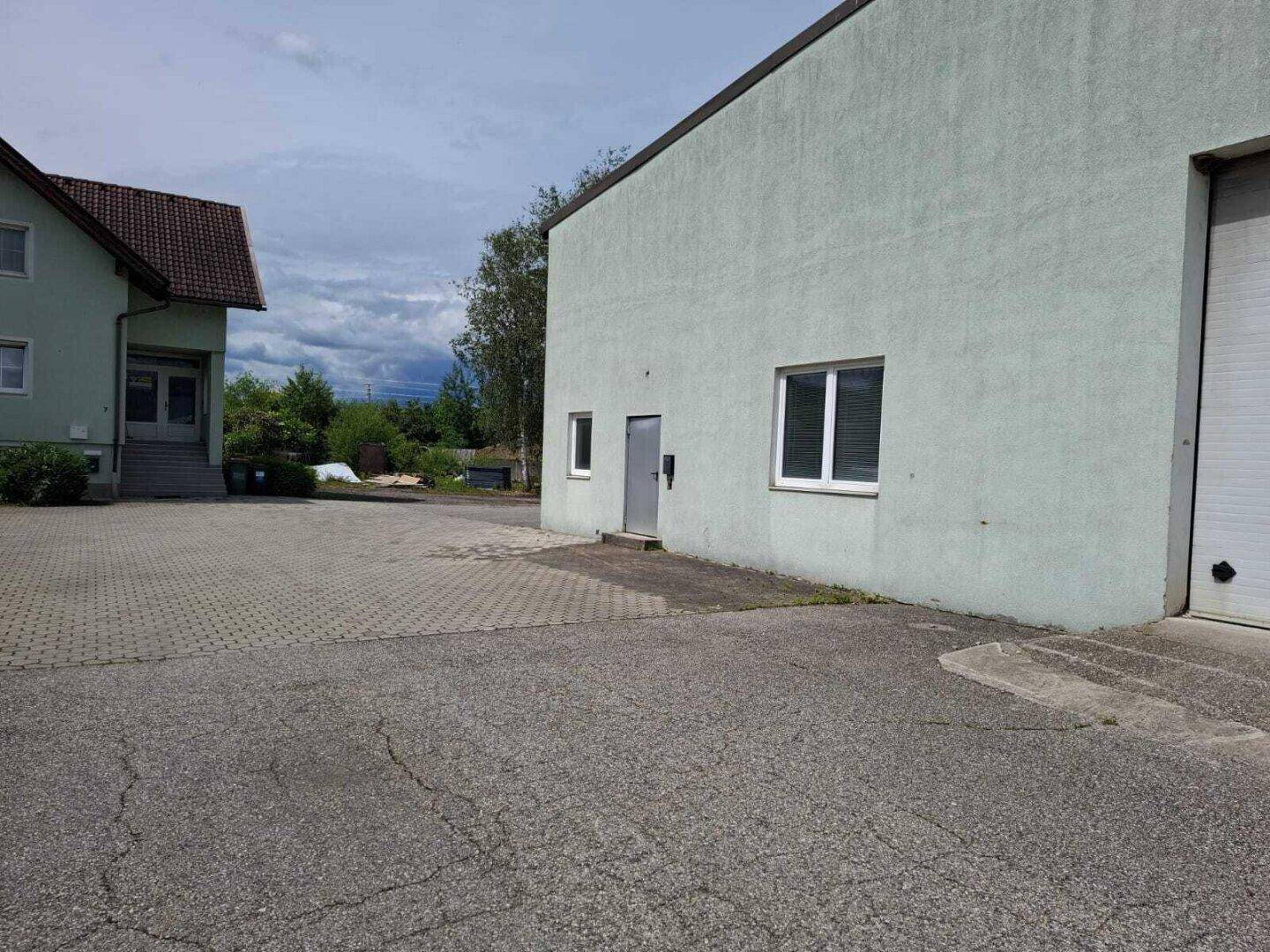 Außenbereich mit zwei Gebäuden, einem Industriegebäude mit Rolltor und einem kleineren Wohngebäude, sowie gepflastertem Vorplatz.