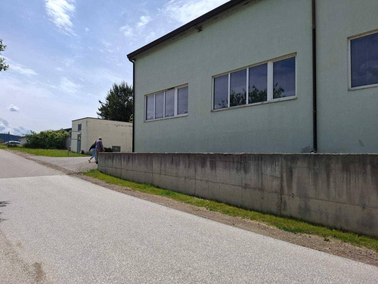 Seitenansicht des hellgrünen Industriegebäudes mit einer langen Betonmauer und einer Straße im Vordergrund.