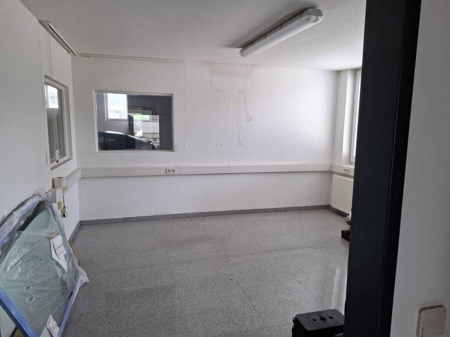Leeres Büro oder kleiner Raum mit weißen Wänden, grauen Fliesen und einem Fenster, das Tageslicht hereinlässt.