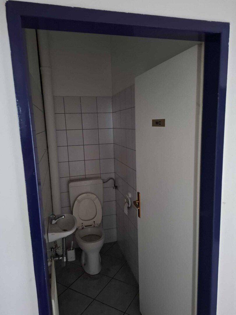 Kleiner Toilettenraum mit weißen Fliesen, einer Toilette und einem Waschbecken, durch eine offene Tür sichtbar.
