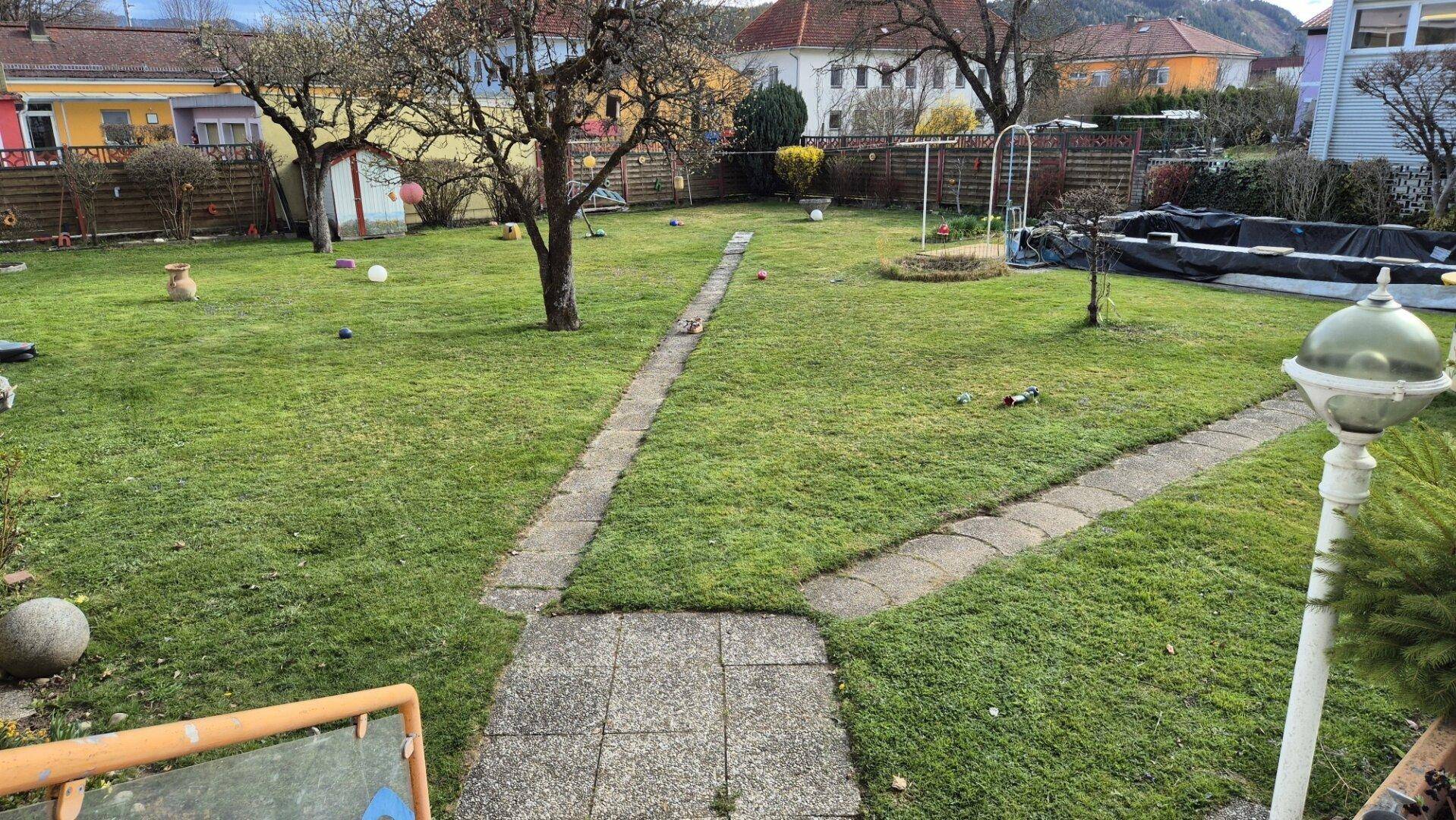 Großer Garten mit Rasenfläche, Bäumen und einem Steinweg, umgeben von Wohngebäuden.