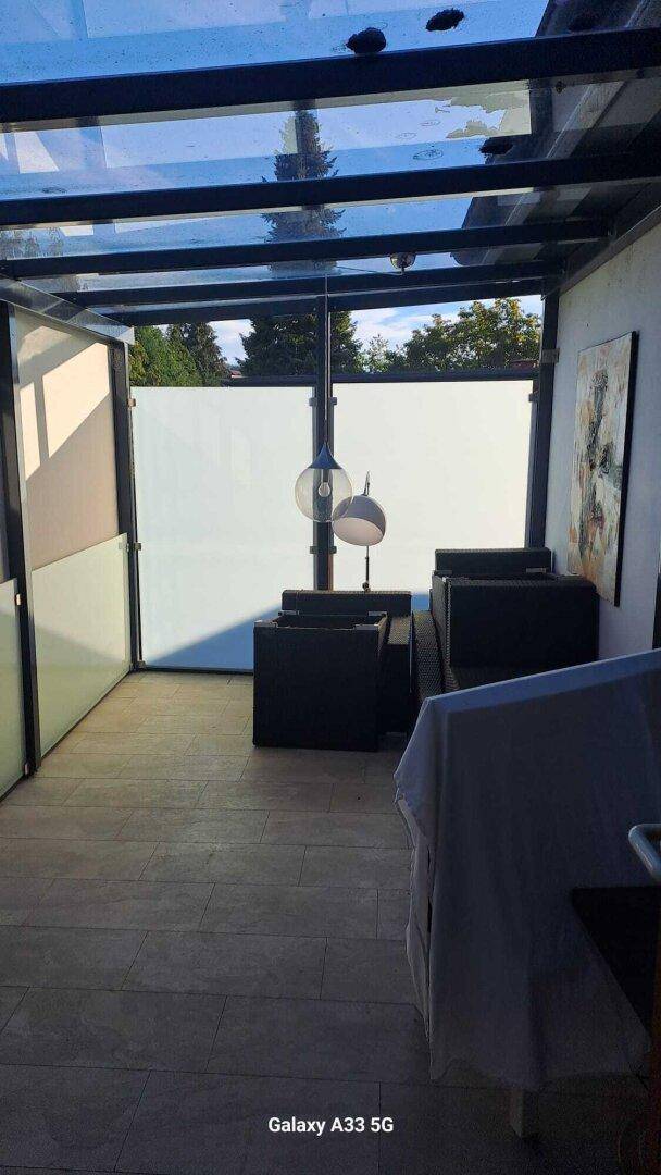 Überdachte Loggia mit Sitzmöbeln, Fliesenboden und Blick ins Grüne.
