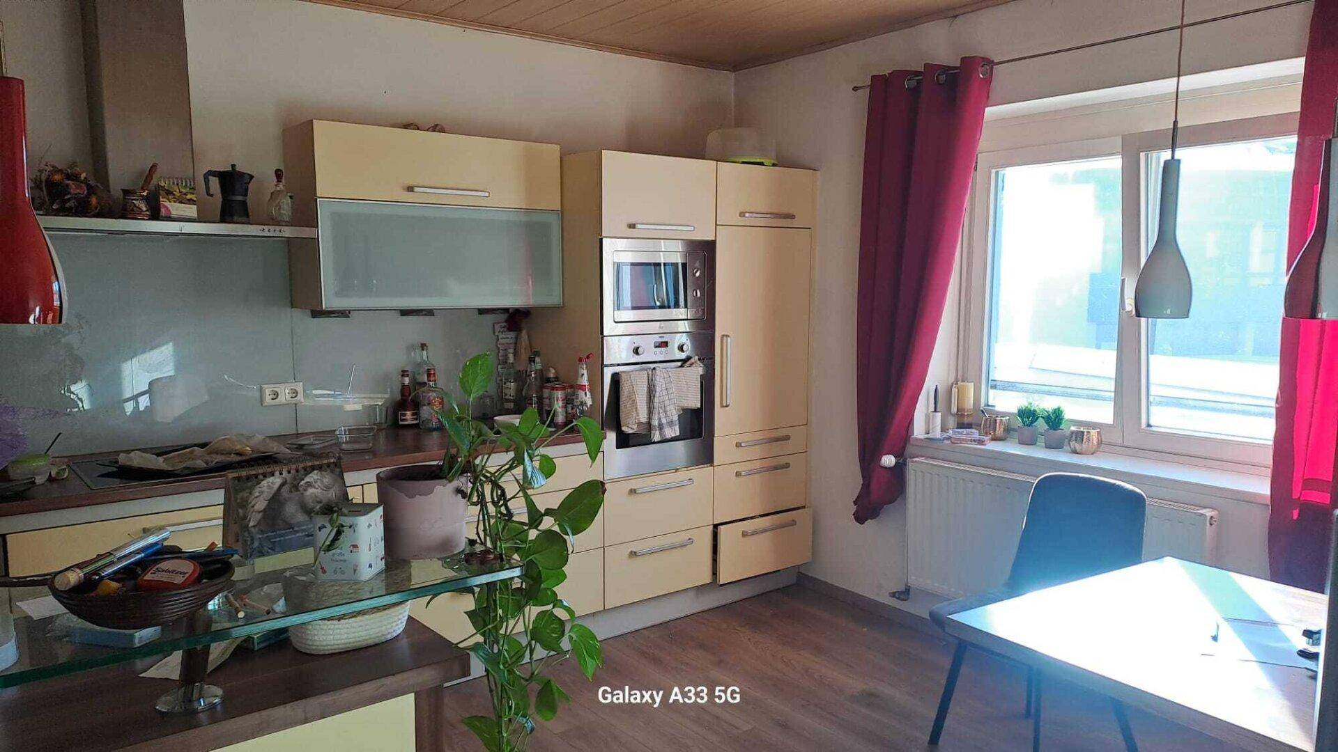 Helle Küche mit beigen Schränken, integriertem Backofen und Essbereich am Fenster.