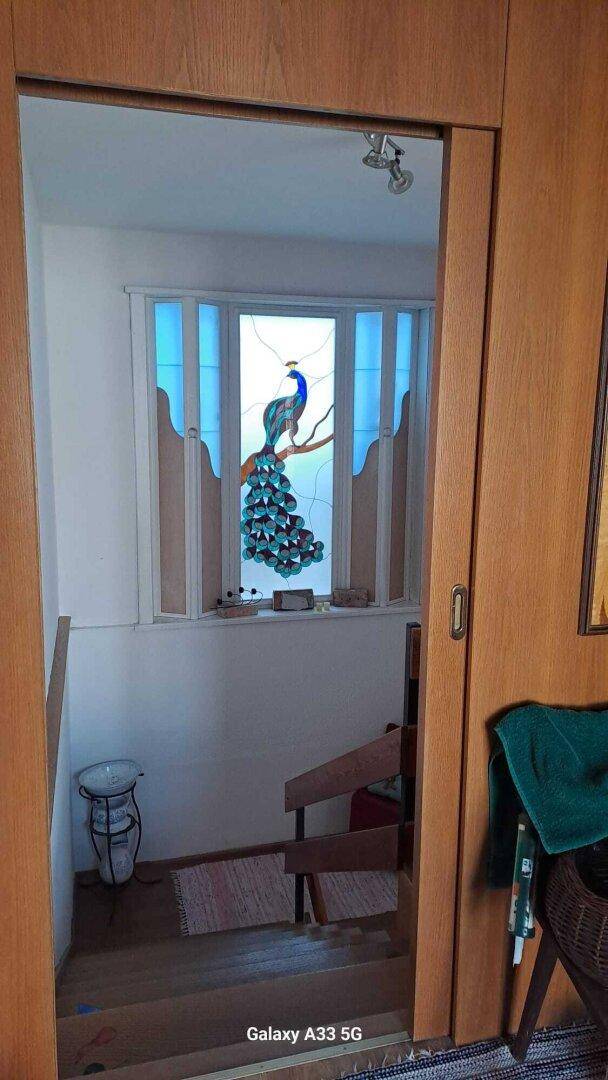 Treppenbereich mit Buntglasfenster, das einen Pfau darstellt, und Holztreppe.