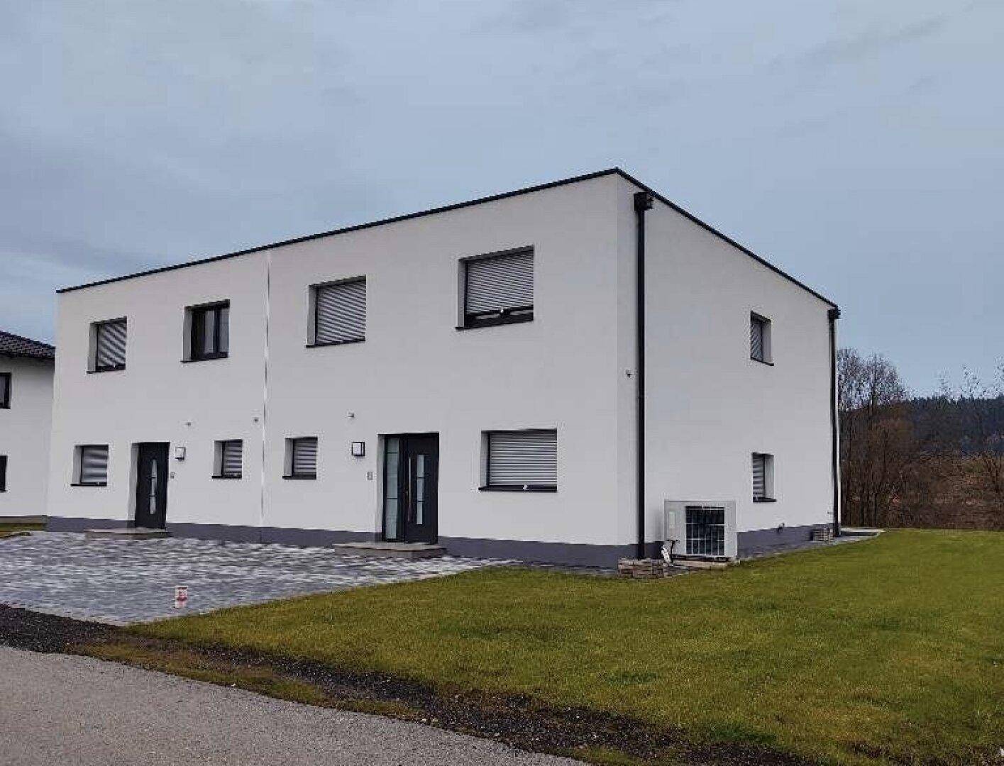 Moderne Doppelhaushälfte mit weißer Fassade, grauen Akzenten und Rollläden unter bewölktem Himmel.