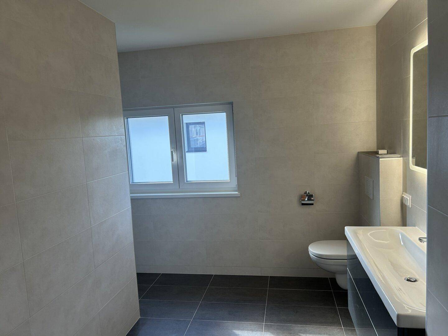 Modernes Badezimmer mit Fliesen, beleuchtetem Spiegel und Fenster, das eine helle Atmosphäre schafft.