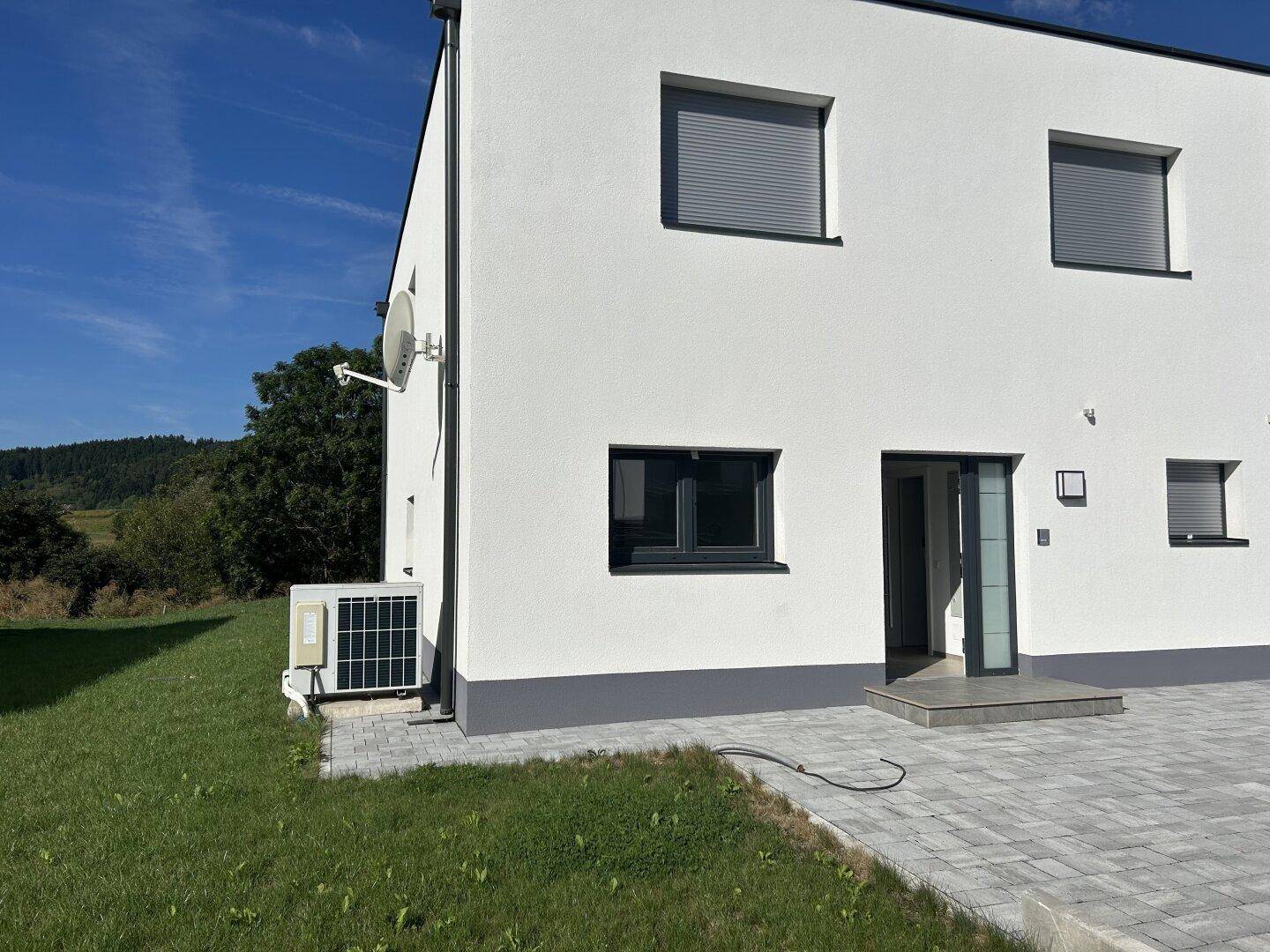 Seitenansicht einer modernen Doppelhaushälfte mit Klimaanlage, gepflastertem Bereich und grüner Umgebung.