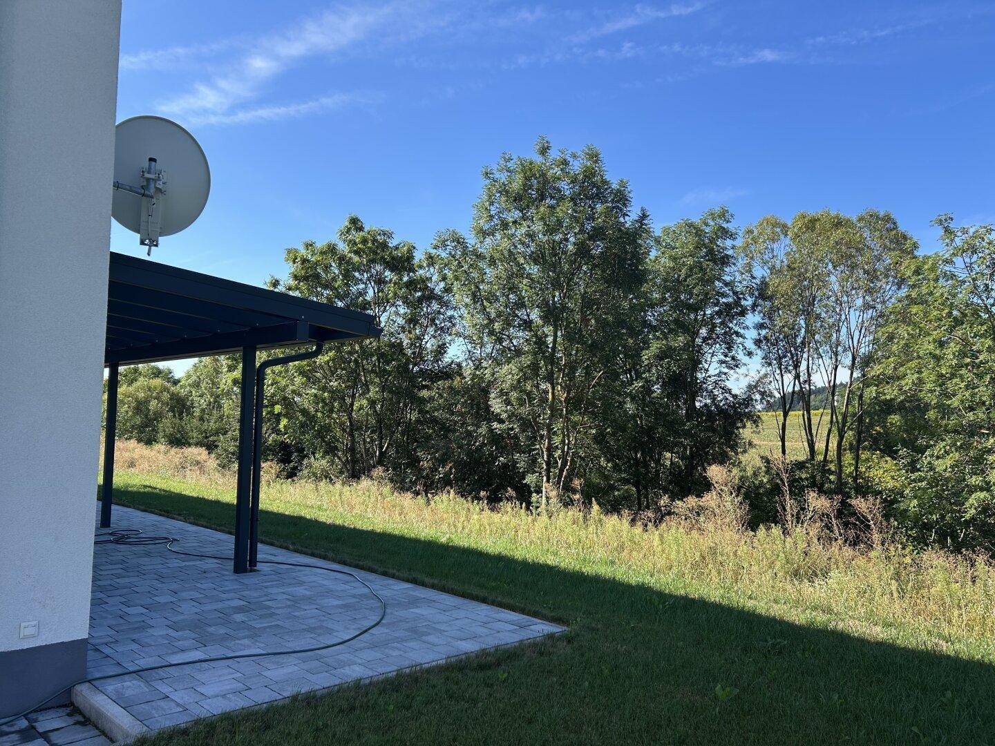 Gepflasterte Terrasse mit Überdachung und weitem Blick in die grüne Landschaft unter blauem Himmel.