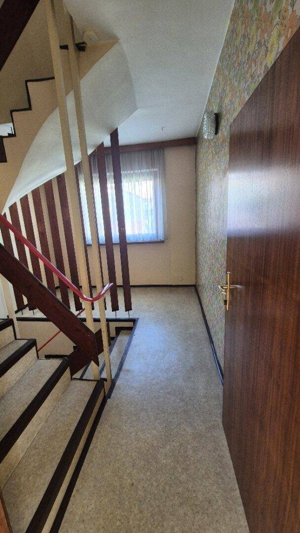 Treppenhaus mit Teppichboden, Holzgeländer und Fenster, das Tageslicht hereinlässt.