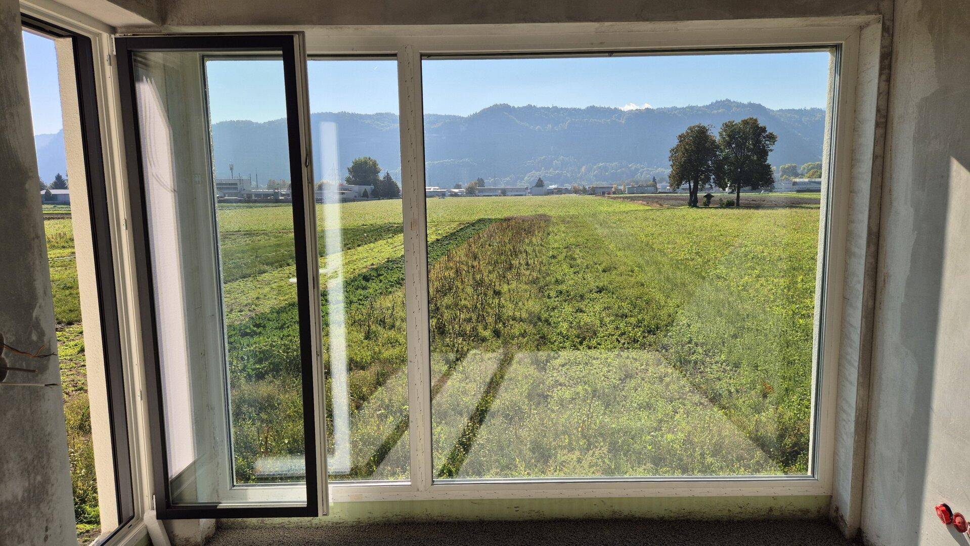 Unfertiger Innenraum mit großen Fenstern und weitem Blick auf grüne Felder und Berge.