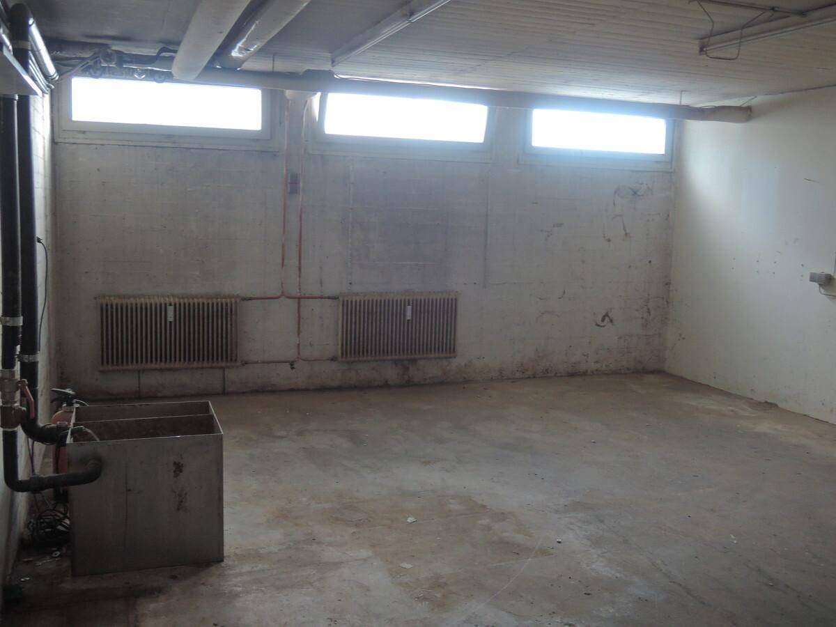 Leerer Kellerraum mit kleinen Fenstern, Betonboden und sichtbaren Heizkörpern an den Wänden.