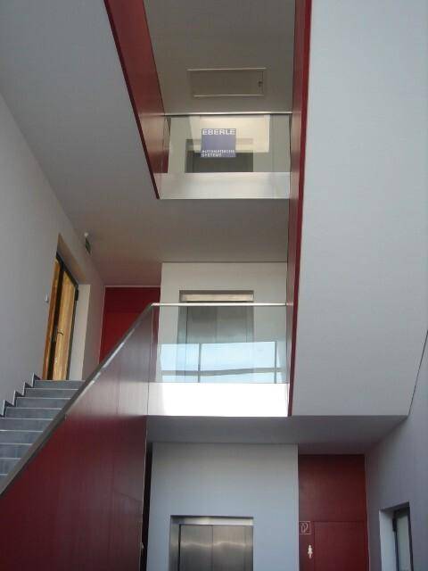 Helles Treppenhaus mit moderner Architektur, Glasgeländern, roten Wänden und einem Aufzug im unteren Bereich.