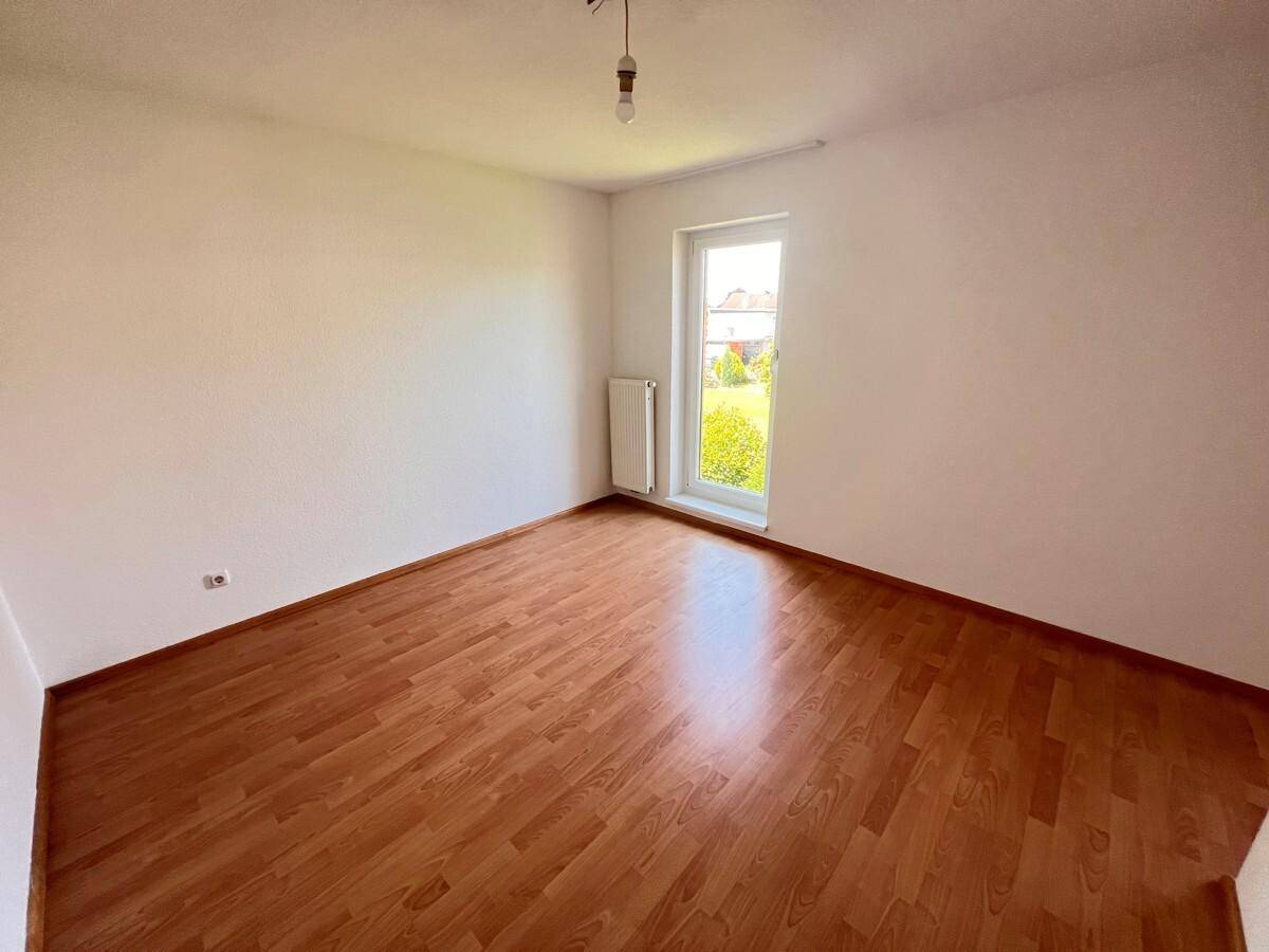 Leeres Schlafzimmer mit Holzboden und einem Fenster mit Blick ins Grüne.
