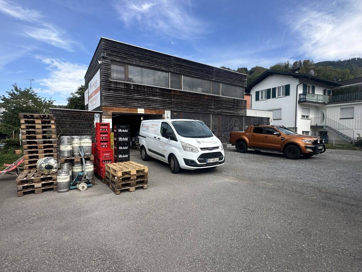 Gewerbegebäude mit Holzfassade, davor geparkte Fahrzeuge und gestapelte Paletten auf einem Schotterparkplatz.