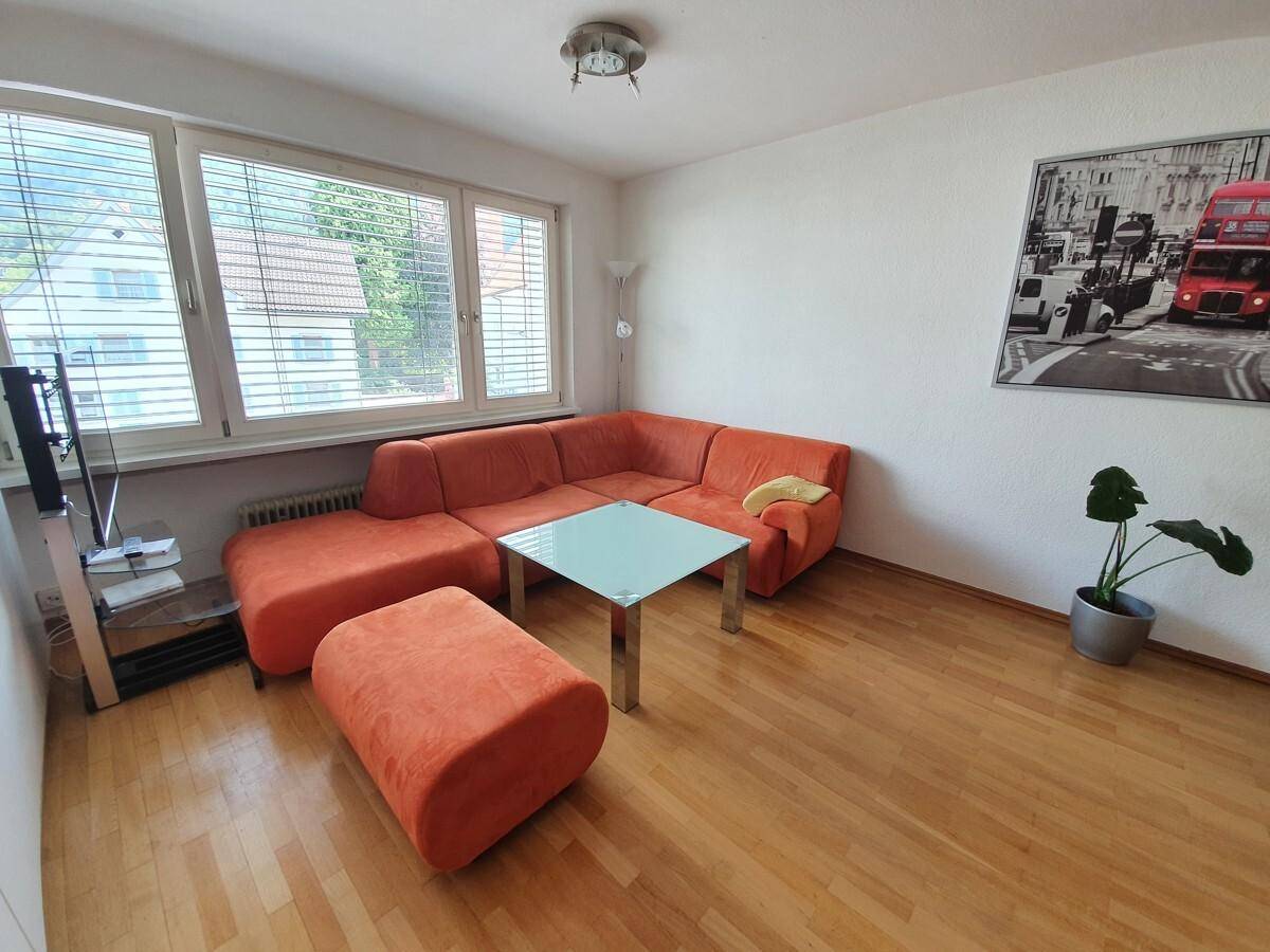 Gemütliches Wohnzimmer mit orangefarbener Eckcouch, Holzboden und großem Fenster mit Jalousien.
