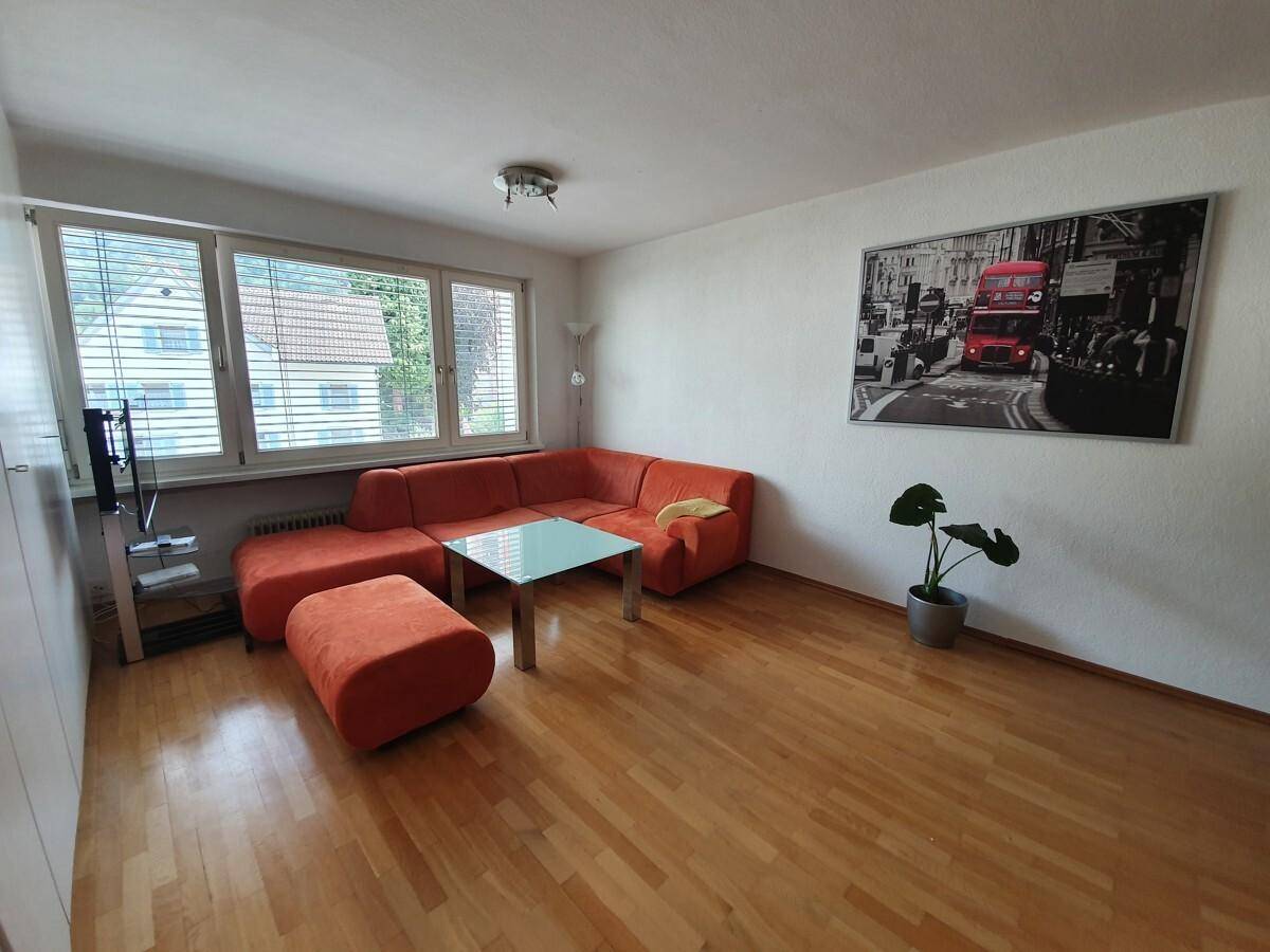 Lichtdurchflutetes Wohnzimmer mit bequemer orangefarbener Couch, Holzboden und Blick ins Grüne.