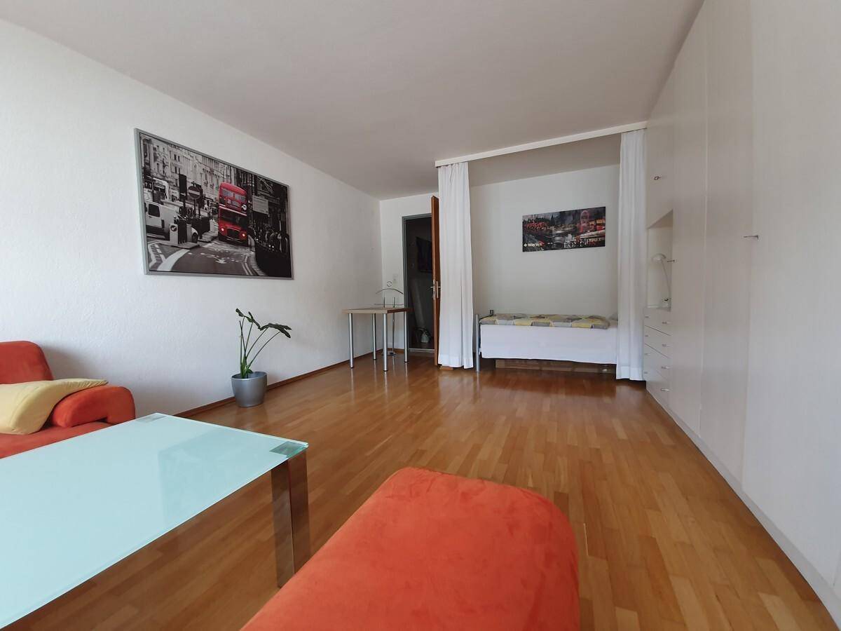 Geräumiges Wohnzimmer mit Holzboden, weißem Wandschrank und abgetrenntem Schlafbereich im Hintergrund.