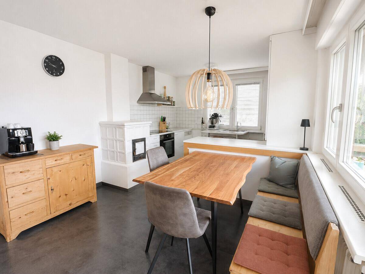 Helle Wohnküche mit modernem Essbereich, Holztisch und traditionellem Kachelofen.
