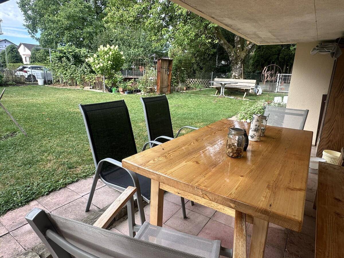 Gepflasterte Terrasse mit Holztisch und Stühlen, angrenzend an einen grünen Garten.