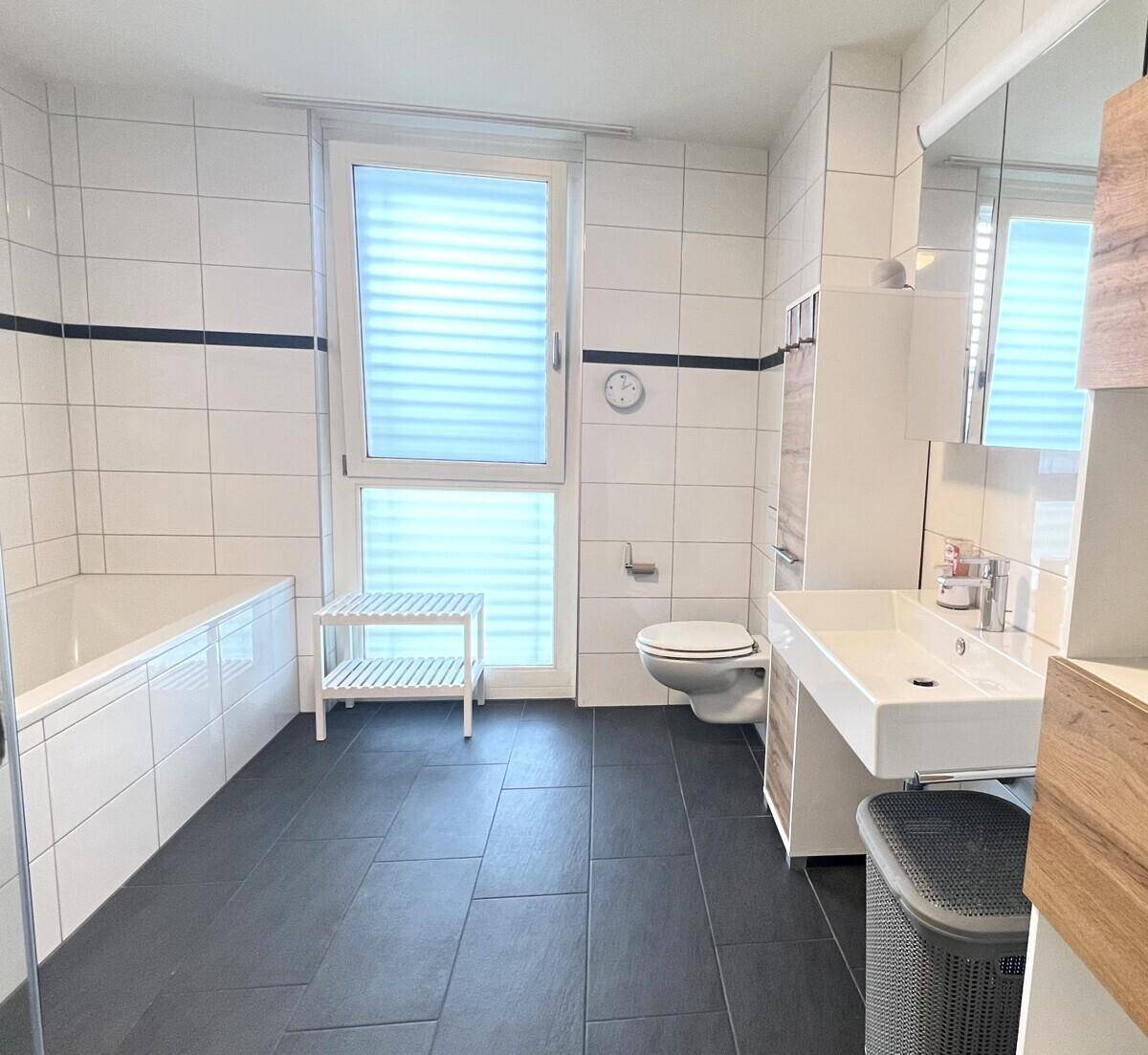Modernes Badezimmer mit Badewanne, Waschtisch, WC und Fenster, ausgestattet mit dunklen Bodenfliesen.