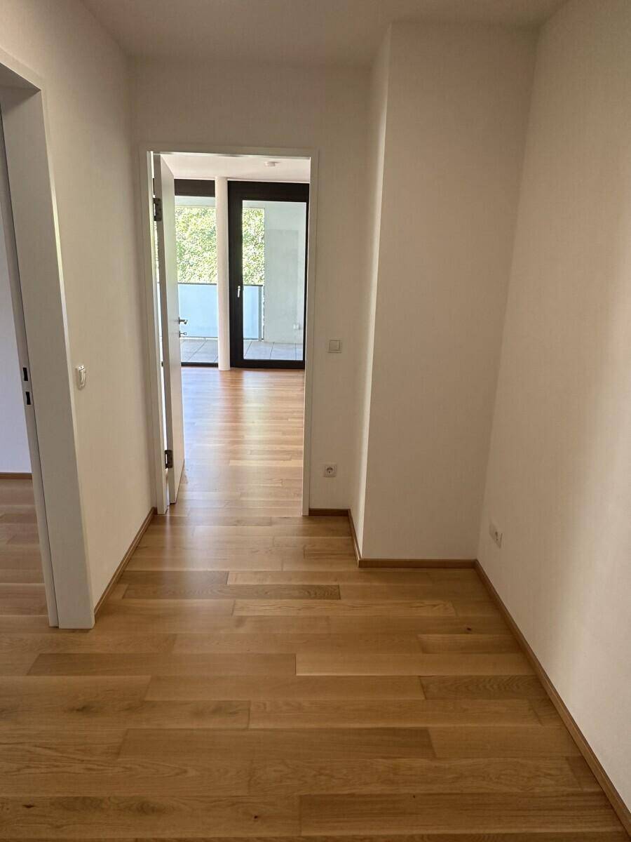 Heller Flur mit Holzboden, der zu den Zimmern und einem Balkon führt.