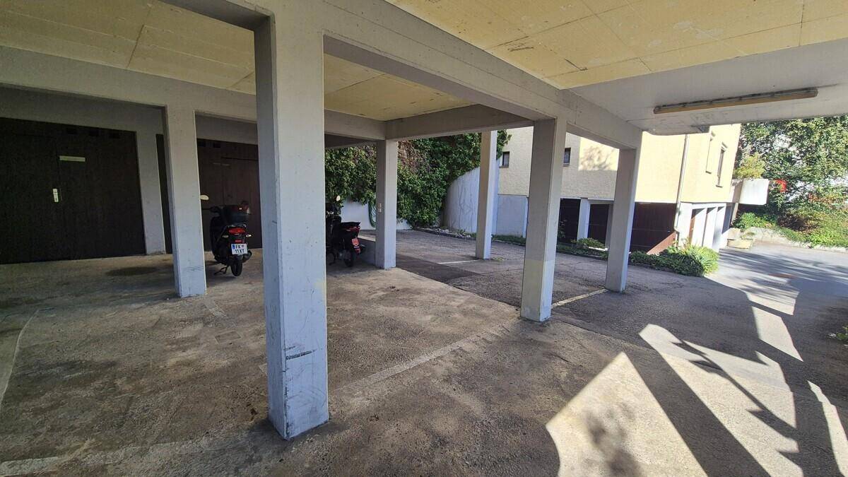 Überdachte Parkfläche mit mehreren Stellplätzen, Betonpfeilern und angrenzendem Gebäude.