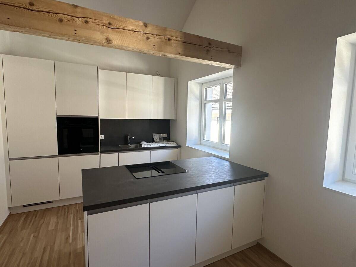 Moderne Einbauküche mit weißen Schränken, dunkler Arbeitsplatte und Kochinsel unter Holzbalken.