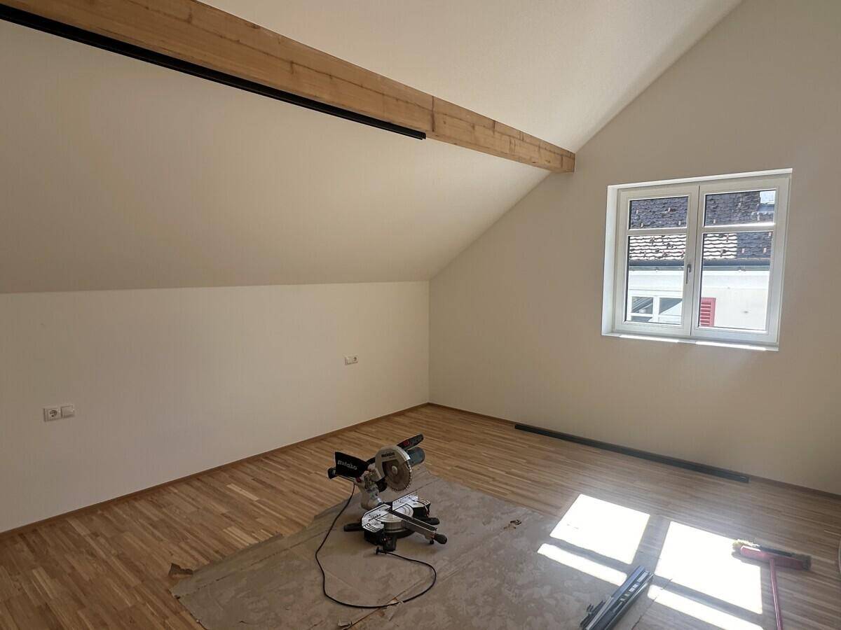 Dachbodenraum mit Holzbalken, Schrägdach und Fenster, bereit für den Innenausbau.