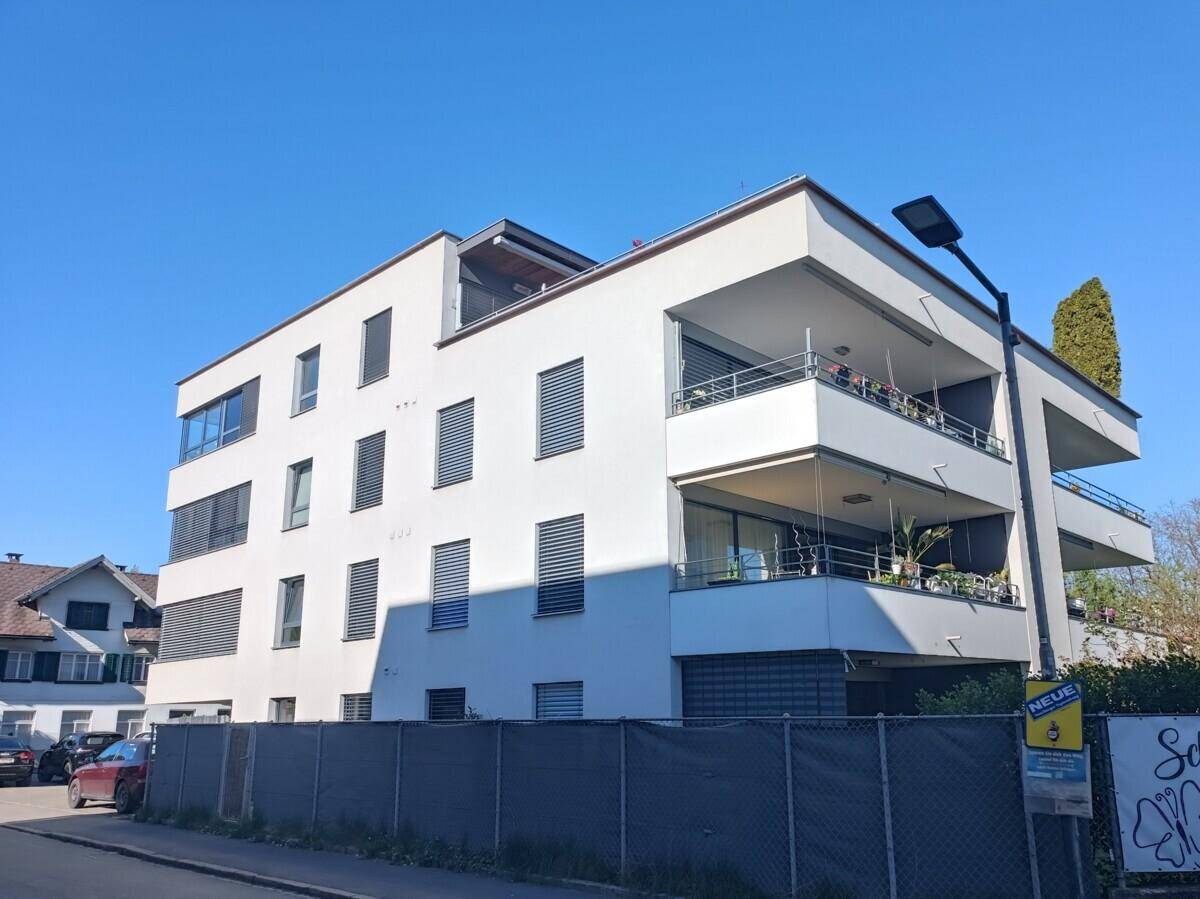 Modernes Mehrfamilienhaus mit weißer Fassade, Balkonen und Jalousien unter blauem Himmel.