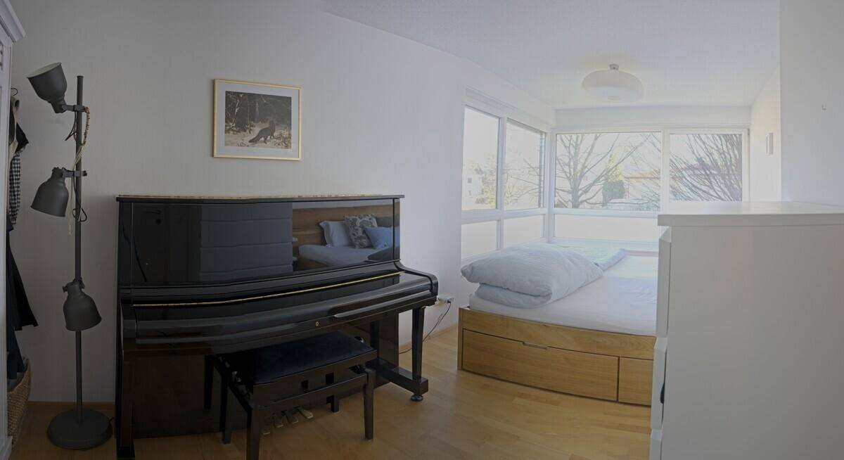 Schlafzimmer mit Holzboden, einem Klavier und großen Fenstern, die viel Licht hereinlassen.