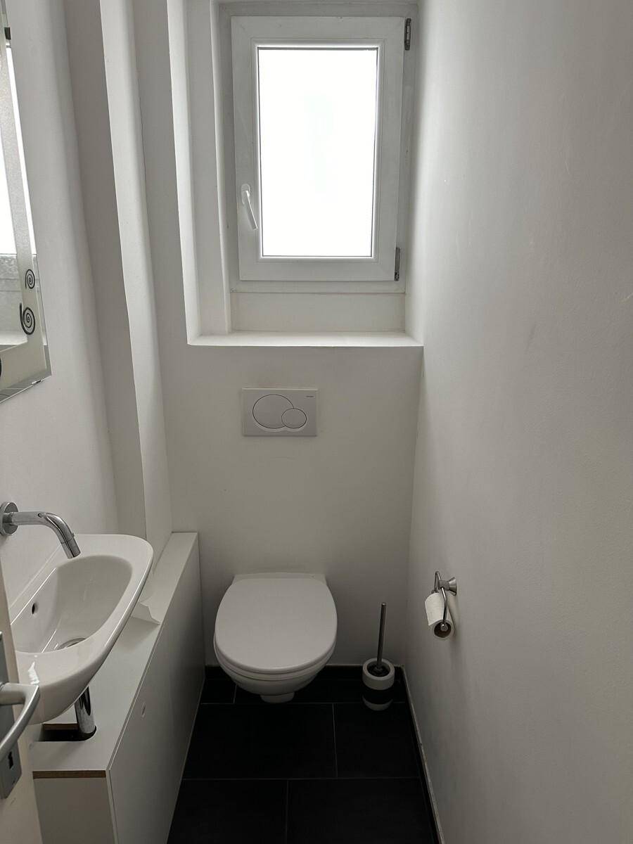 Kleines Gäste-WC mit wandhängender Toilette, kleinem Waschbecken und Fenster für Tageslicht.