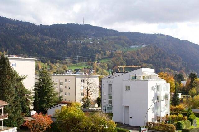 Panoramablick auf Wohngebäude und eine grüne Hügellandschaft mit Bergen im Hintergrund.