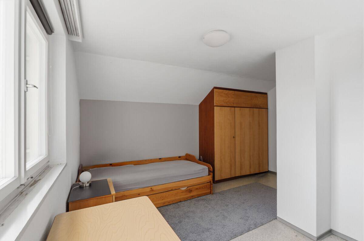 Einfaches Schlafzimmer mit Einzelbett, Holzschrank und Fenster, das den Raum erhellt.