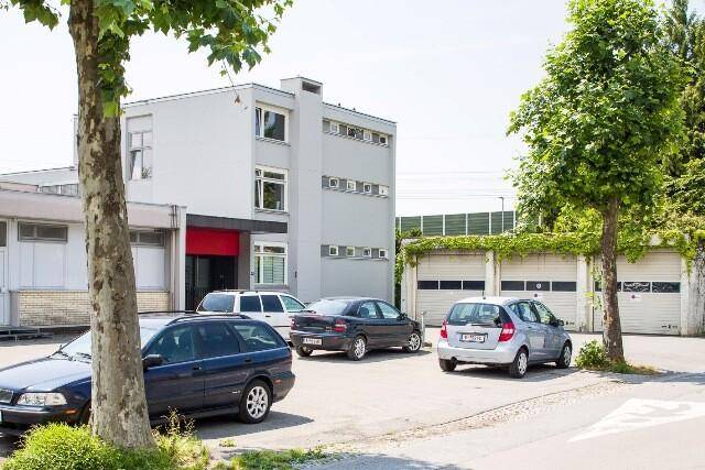 Außenansicht eines Gewerbegebäudes mit Parkplätzen und Bäumen, umgeben von einer grünen Landschaft.