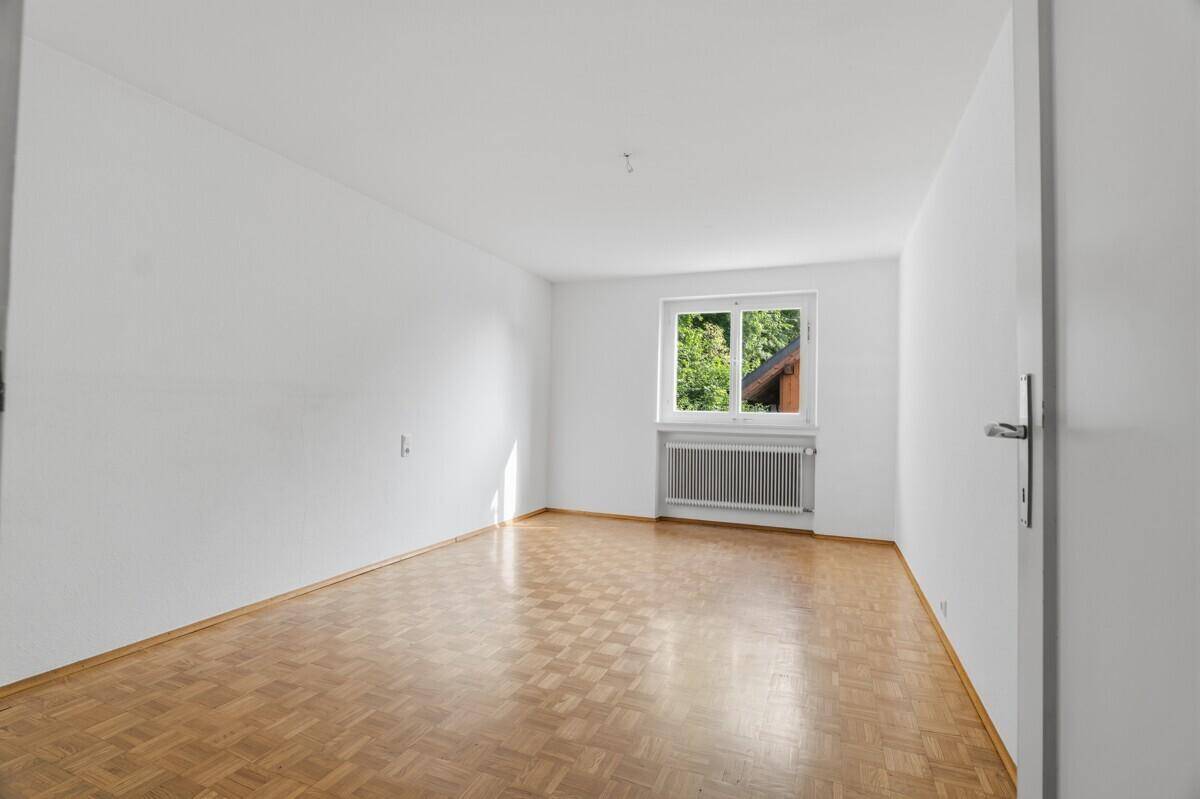 Leerer Raum mit Parkettboden und einem Fenster, das einen Blick ins Grüne bietet.