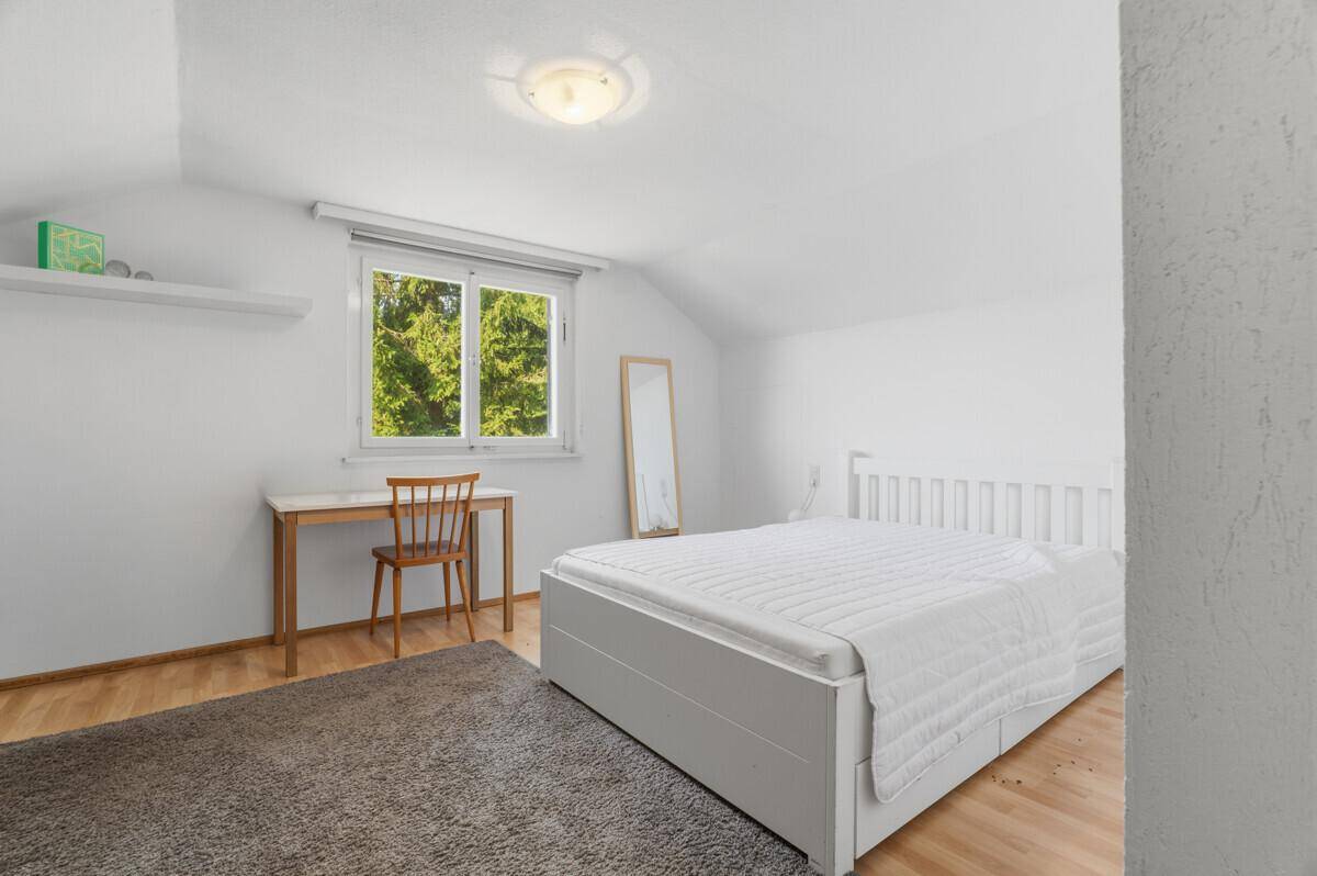 Helles Schlafzimmer mit Einzelbett, Holzschreibtisch und Blick ins Grüne durch das Fenster.